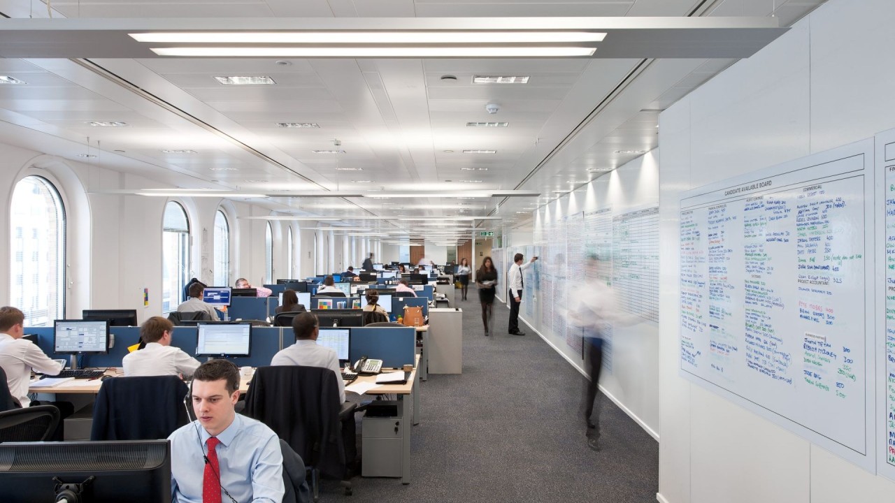 1745428842356 Open-plan office with multiple people working at desks with computers; some people are sitting, while others stand or walk by a large wall covered with notes and schedules. Bright, modern environment with large windows.