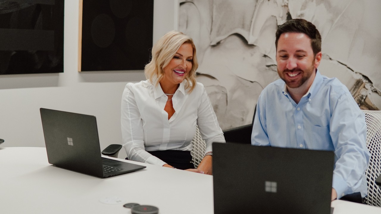 1745860601328 Two colleagues, a woman and a man, sit at a white table with laptops in a modern office, smiling as they look at the screen. Abstract artwork hangs on the wall behind them.