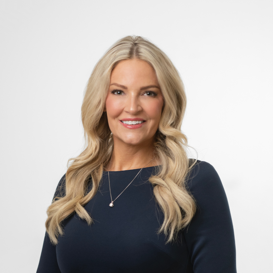 A woman with long, wavy blonde hair wearing a navy blue top and a delicate necklace smiles at the camera against a plain white background.