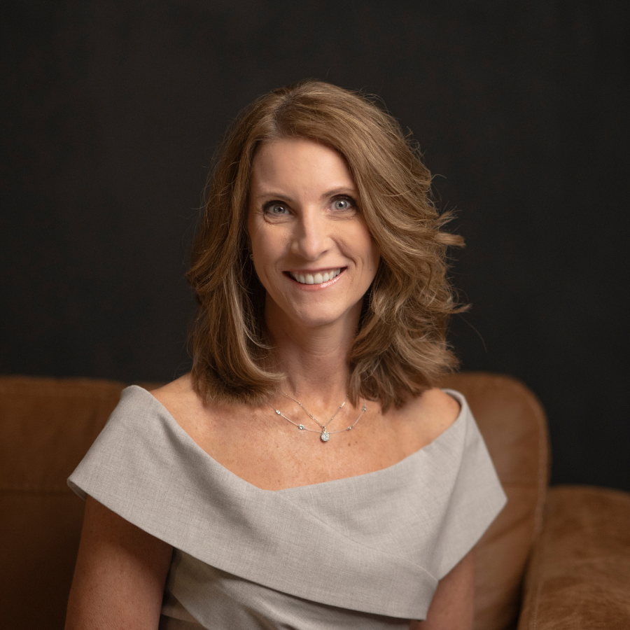 A woman with shoulder-length light brown hair and blue eyes, wearing a light gray off-the-shoulder dress and a necklace, smiles while sitting on a brown couch against a dark background.