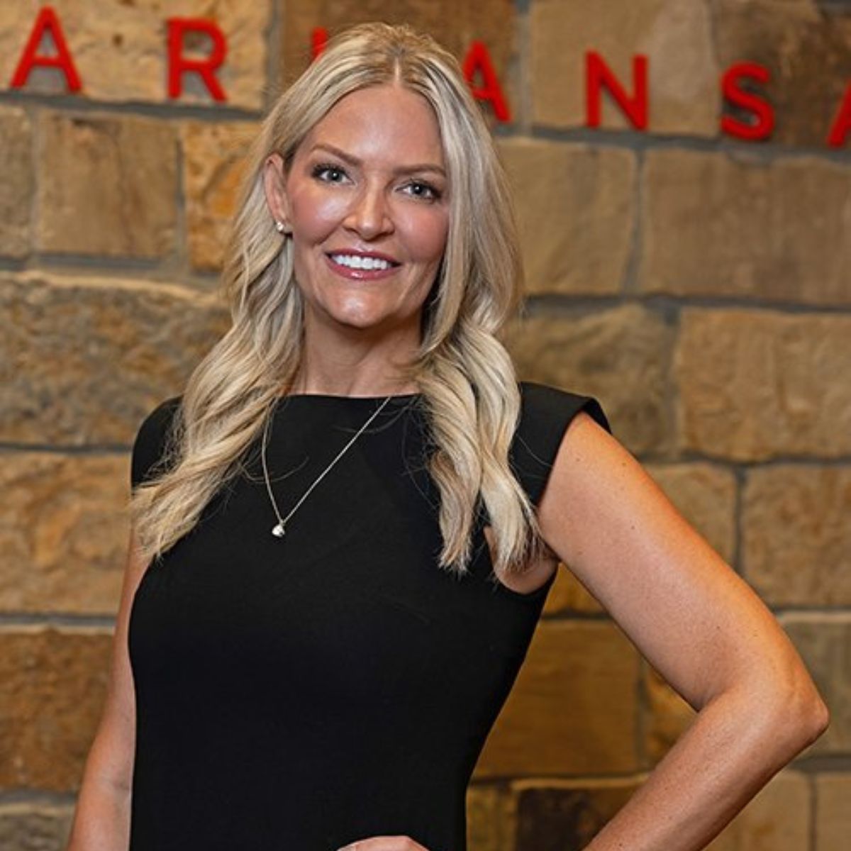A smiling woman with long blonde hair wearing a black sleeveless dress stands in front of a stone wall with red letters partially spelling ARKANSAS in the background.