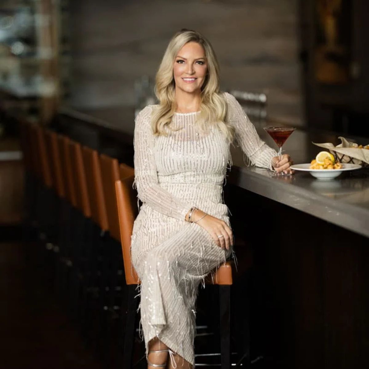 A woman with long blonde hair, wearing a sparkly white dress, sits at a bar holding a martini glass. There is a plate of food next to her, and empty barstools line the counter. She is smiling at the camera.
