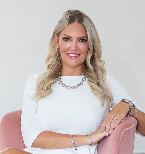 A woman with long blonde hair, wearing a white dress and silver jewelry, sits smiling on a pink chair against a plain background.